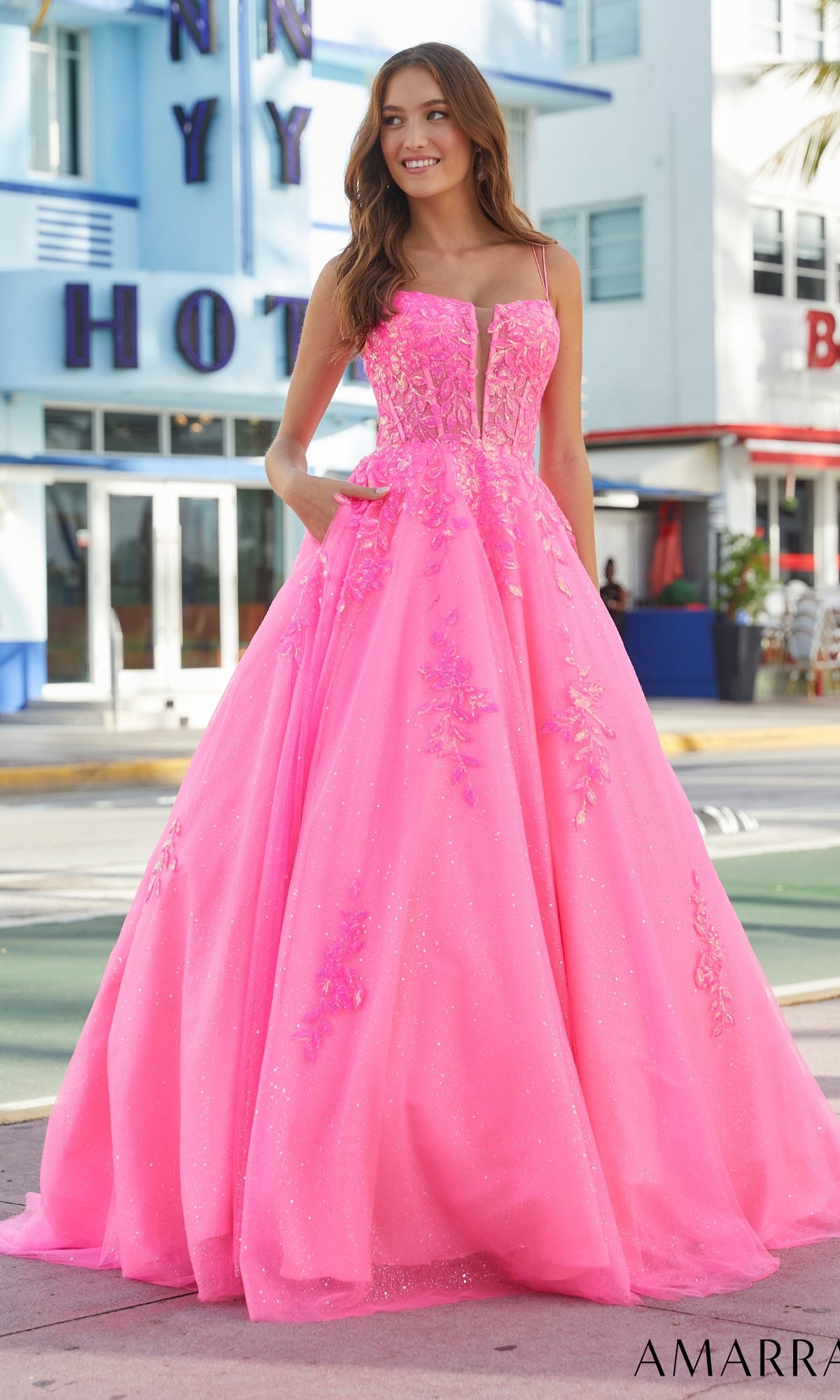 A woman stands outdoors in front of a hotel, wearing the Amarra Long Prom Dress 88574—a bright pink gown with floral details, a glittering tulle skirt, and an elegant corset bodice.